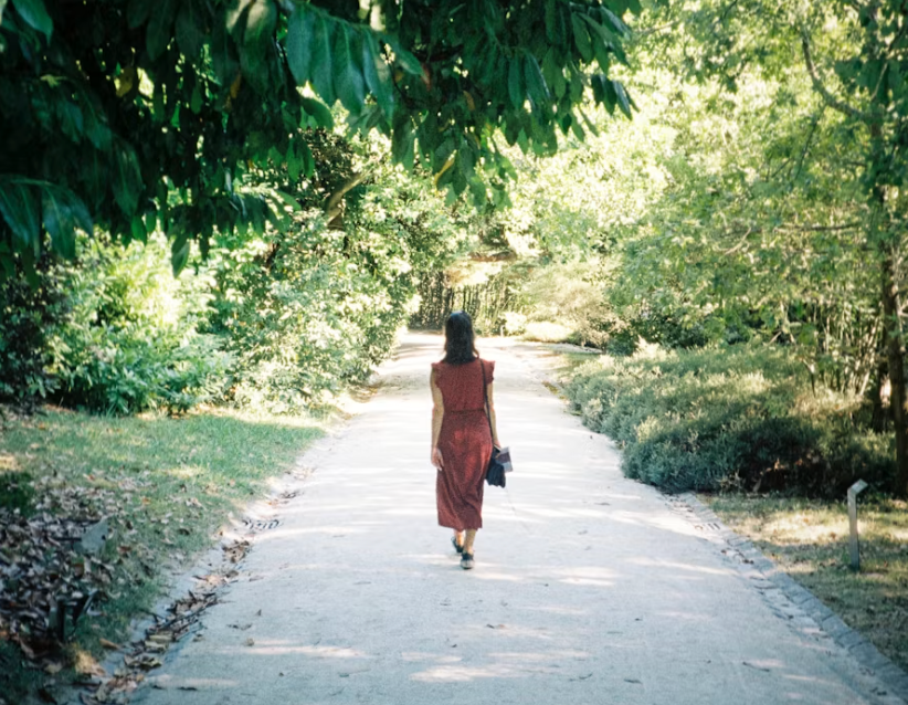 A woman in a rust-coloured dress walks alone down a tree-lined path, lost in thought.
