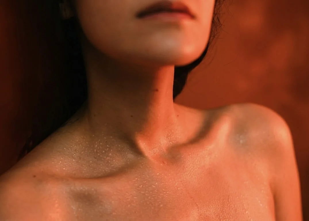 Close-up of a woman's neck and collarbone, skin lightly beaded with sweat in warm low light, evoking a perimenopause night sweat.