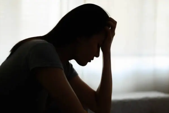 A woman sits in silhouette against a softly lit window, head bowed with one hand to her forehead, conveying low mood and emotional distress.