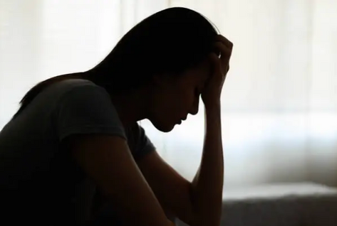 A woman sits in silhouette against a softly lit window, head bowed with one hand to her forehead, conveying low mood and emotional distress.